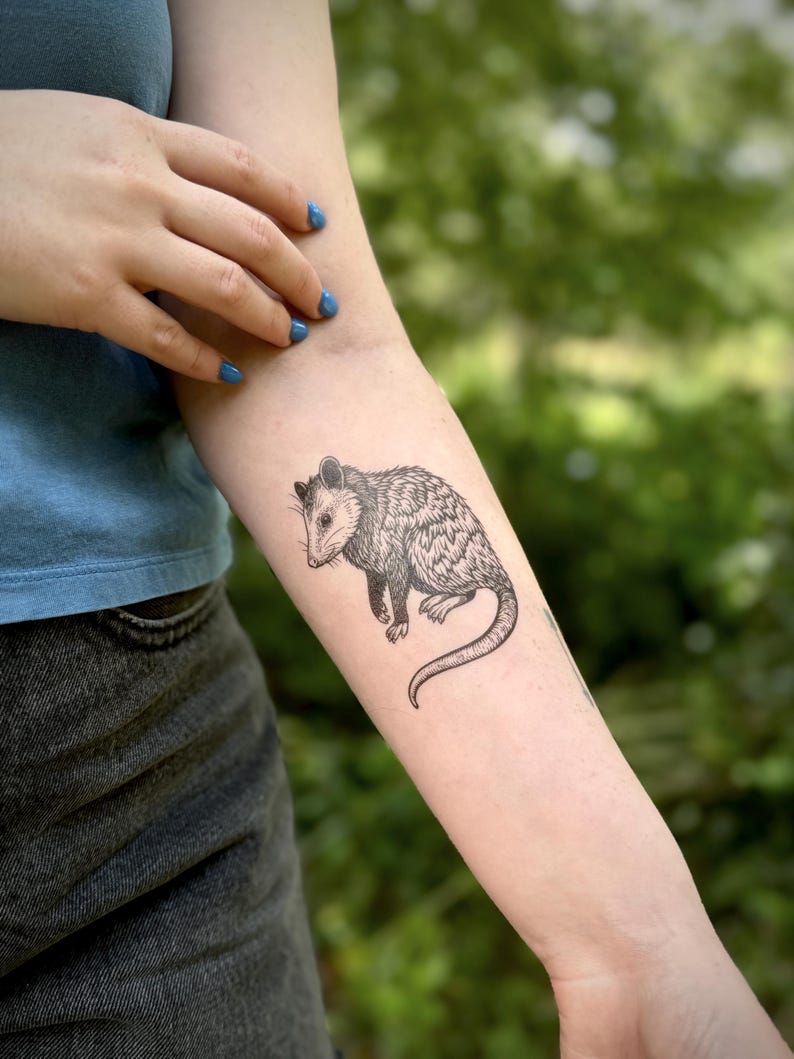 May include: A black and white tattoo on a forearm featuring a detailed illustration of an opossum. The animal is depicted in profile with fine line work showing its fur texture, pointed snout, small ears, and a long, curved, hairless tail.