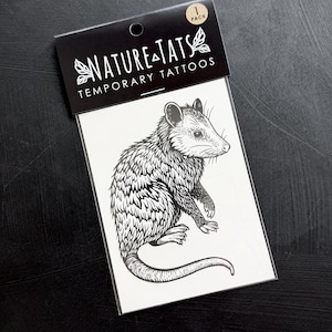 May include: A black and white temporary tattoo sheet featuring a detailed line art illustration of an opossum. The packaging header reads NATURE TATS TEMPORARY TATTOOS and includes a 1 PACK label. The design is printed on a white rectangular backing.