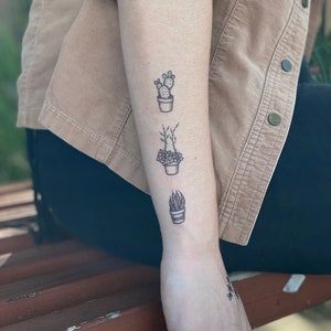 May include: A person's forearm with three small potted plant tattoos. The tattoos are in black ink and feature a cactus, a succulent, and a snake plant.