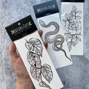 May include: Three temporary tattoo sheets with black line drawings of a snake, a rose, and a monstera leaf. The sheets are labeled "Nature Tats Temporary Tattoos".