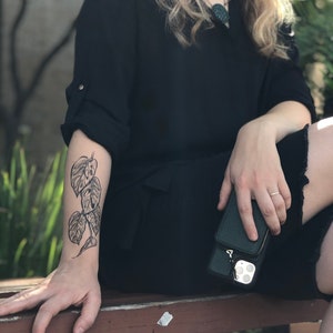 May include: A person's arm with a black line drawing tattoo of a plant with large leaves. The tattoo is on the forearm and is visible from the wrist to the elbow.