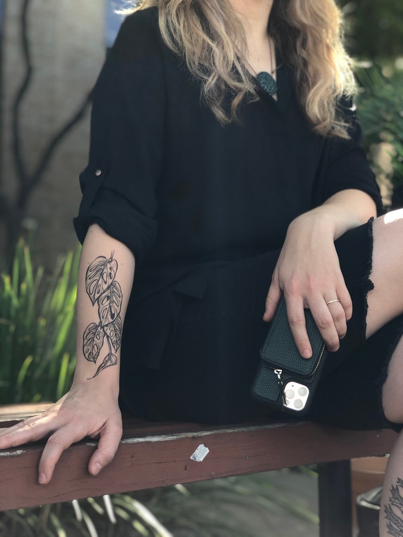 May include: A person's arm with a black line drawing tattoo of a plant with large leaves. The tattoo is on the forearm and is visible from the wrist to the elbow.