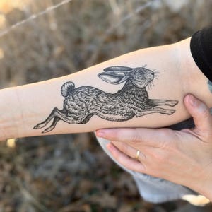 Puede incluir: Ilustración en blanco y negro de una liebre corriendo tatuada en un brazo. La liebre está representada en un estilo vintage detallado, con pelaje texturizado y orejas largas. El tatuaje está sobre piel clara.