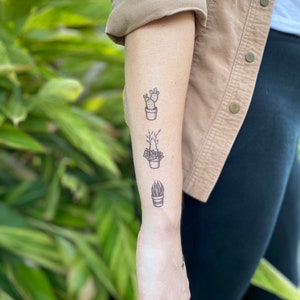 May include: A person's forearm with three small black and white tattoos of potted plants. The tattoos are in a vertical line, with a cactus at the top, a leafy plant in the middle, and a succulent at the bottom.