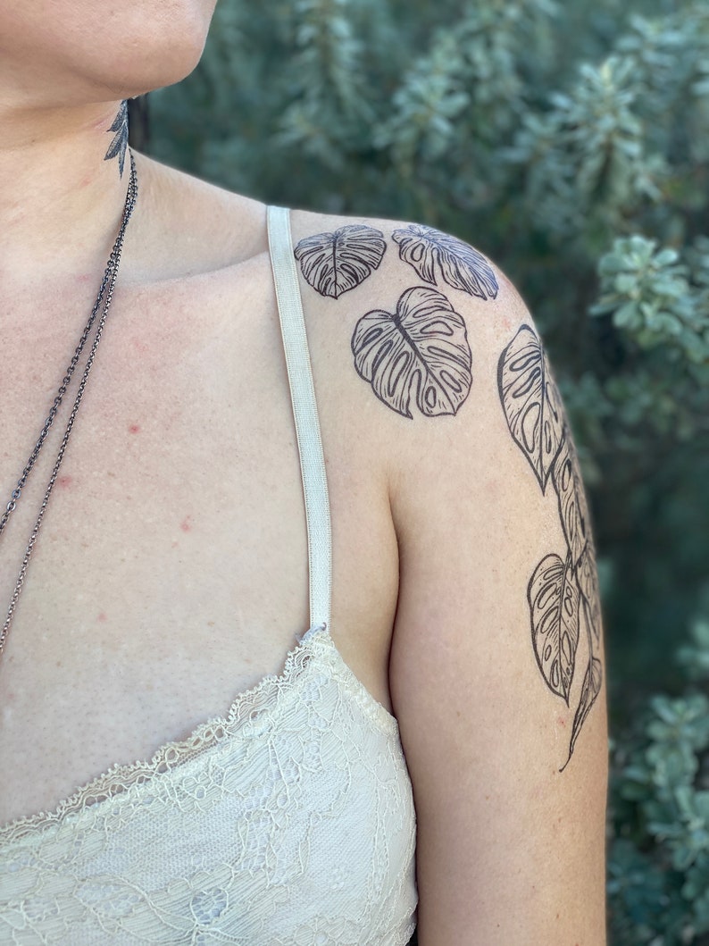 May include: A person's shoulder with a black line drawing tattoo of monstera leaves.