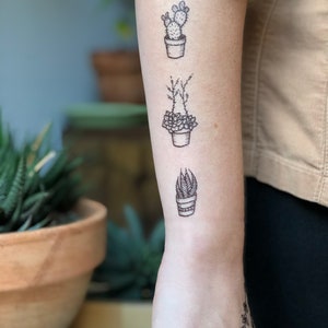 May include: Three black and white temporary tattoos of potted plants on a person's arm. The tattoos feature a cactus, a succulent, and a leafy plant.
