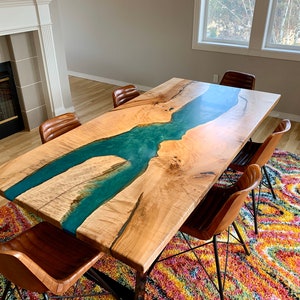 May include: A large wooden dining table with a live edge and a turquoise blue epoxy river running through the center. The table is surrounded by six brown leather chairs with black metal legs.