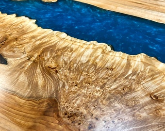 May include: A wooden table top with a blue epoxy river running through the center. The wood has a natural grain pattern and is a light brown color.