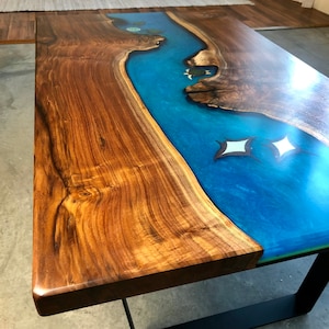 Könnte beinhalten: Eine Tischplatte aus Holz mit einem blauen Epoxid-Fluss, der durch sie verläuft. Das Epoxid hat eine glitzernde Oberfläche und es sind zwei weiße Sterne in das Epoxid eingebettet.