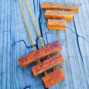 May include: A gold chain necklace with a pendant made of three stacked wooden pieces. The wooden pieces are rectangular and have a reddish-brown color.