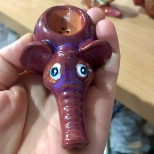 May include: A ceramic elephant pipe with a brown and purple glaze. The pipe has a round bowl and a long, thin spout. The elephant has blue eyes and a small hole in the top of its head.