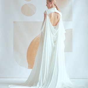 Puede incluir: Un vestido de novia blanco con una cola larga y una espalda baja. El vestido tiene un dise&ntilde;o de capa con un escote drapeado.