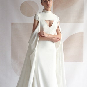 Puede incluir: Un vestido de novia blanco con cuello alto y una larga cola. El vestido tiene un corpi&ntilde;o ajustado y una falda fluida. La modelo est&aacute; de pie frente a un fondo blanco con formas abstractas.