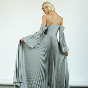 May include: A silver pleated maxi dress with off-the-shoulder sleeves. The dress has a fitted bodice and a flowing skirt.