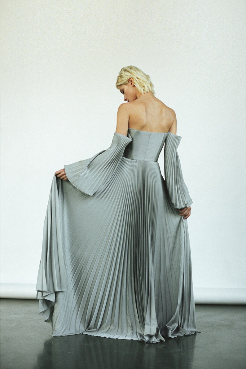 May include: A silver pleated maxi dress with off-the-shoulder sleeves. The dress has a fitted bodice and a flowing skirt.