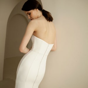 Puede incluir: Un vestido de novia blanco sin tirantes con un corpi&ntilde;o ajustado y una falda fluida. El vestido presenta una fila de botones en la espalda.
