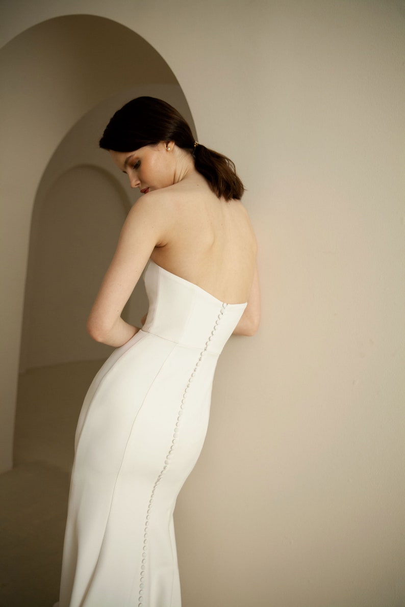 Puede incluir: Un vestido de novia blanco sin tirantes con un corpi&ntilde;o ajustado y una falda fluida. El vestido presenta una fila de botones en la espalda.