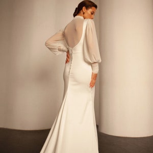 May include: A white wedding dress with a fitted bodice and a flowing skirt. The dress has a sheer, long-sleeved overlay with puffed sleeves. The back of the dress features a low back with a row of buttons.