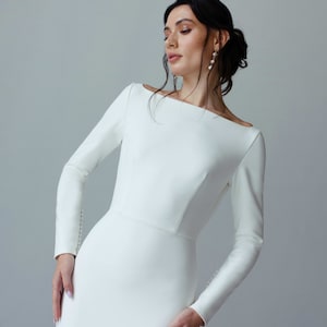 May include: A woman wearing a white, long-sleeved, fitted dress with a bateau neckline. The dress has a simple, elegant design.
