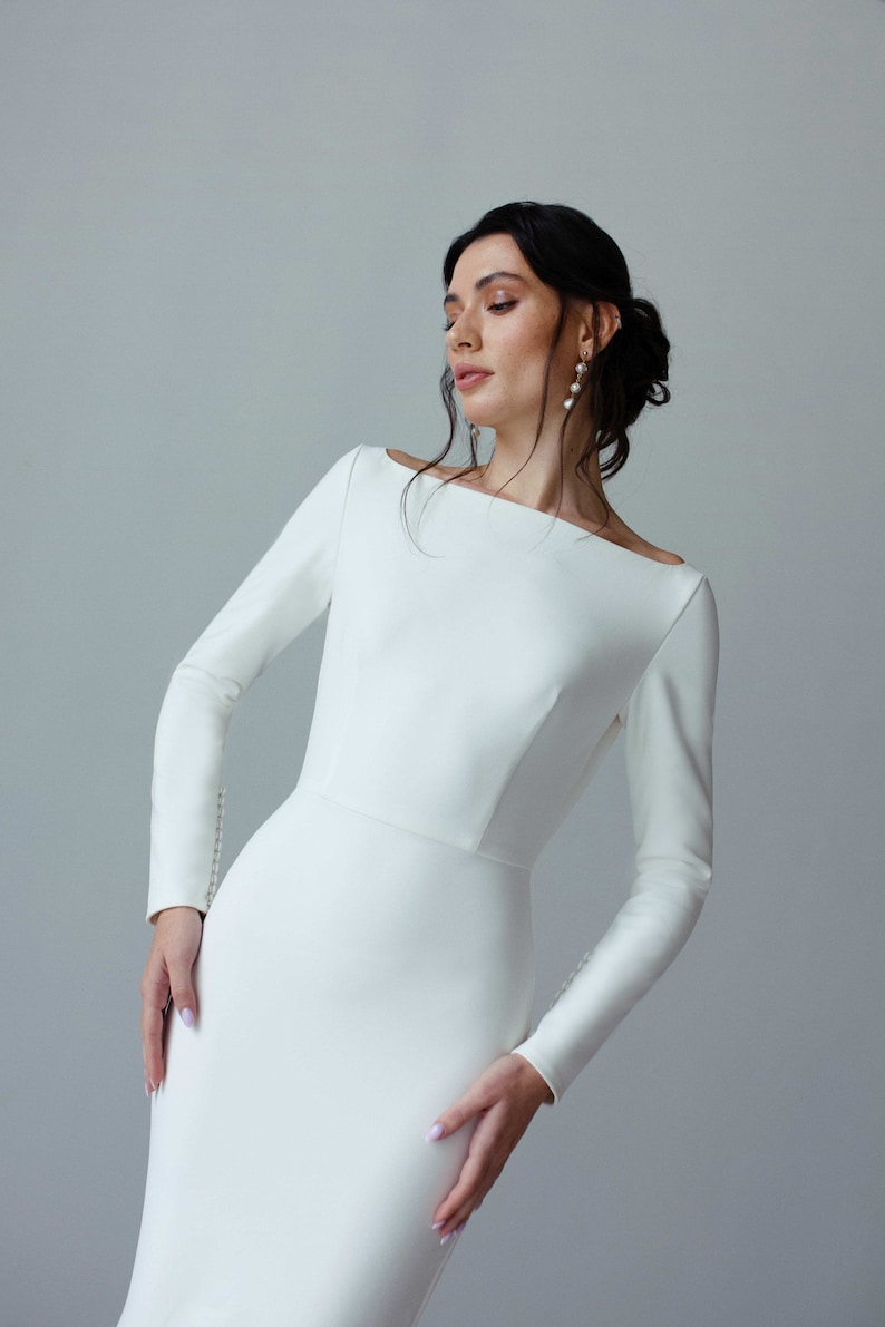 May include: A woman wearing a white, long-sleeved, fitted dress with a bateau neckline. The dress has a simple, elegant design.