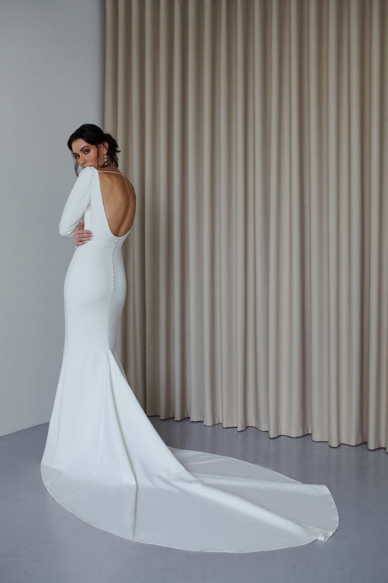 May include: A white, long-sleeved wedding dress with a low back and a train. The dress is fitted and has a mermaid silhouette.