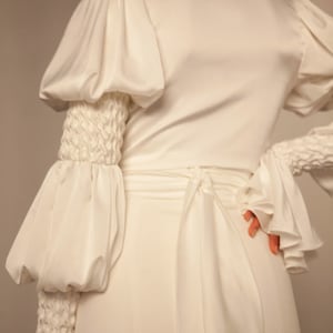 May include: A white satin dress with long sleeves and a cinched waist. The sleeves have large, puffed cuffs and a textured detail at the wrist.