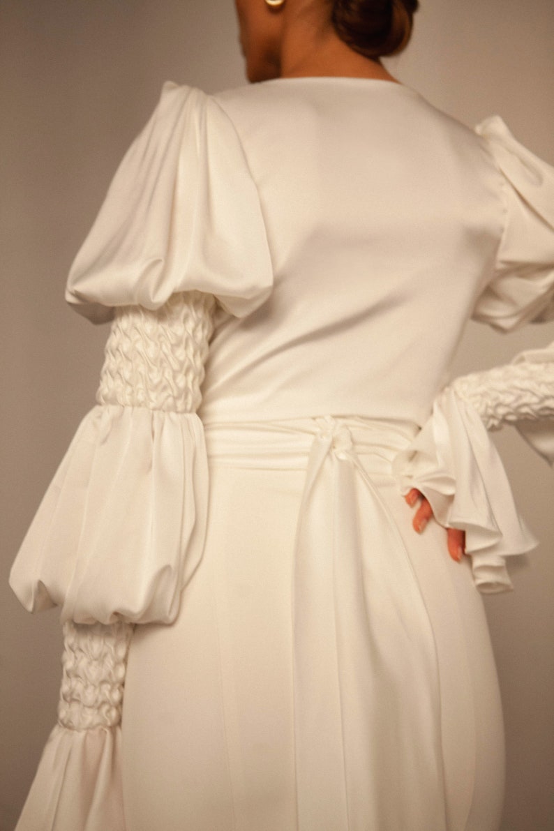 May include: A white satin dress with long sleeves and a cinched waist. The sleeves have large, puffed cuffs and a textured detail at the wrist.