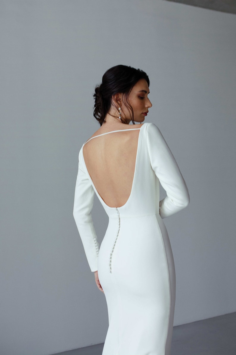 May include: A white long-sleeved wedding dress with a low back and a row of buttons down the back.