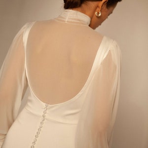 May include: A white wedding dress with a sheer overlay and long sleeves. The dress has a low back with a row of buttons down the centre.