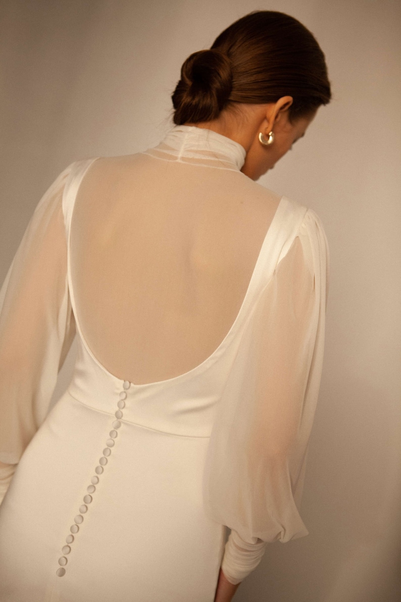 May include: A white wedding dress with a sheer overlay and long sleeves. The dress has a low back with a row of buttons down the centre.