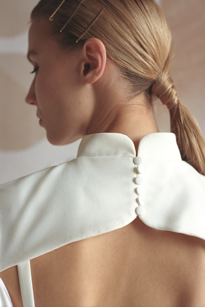 Puede incluir: Una blusa blanca con cuello alto y una fila de botones blancos en la espalda.