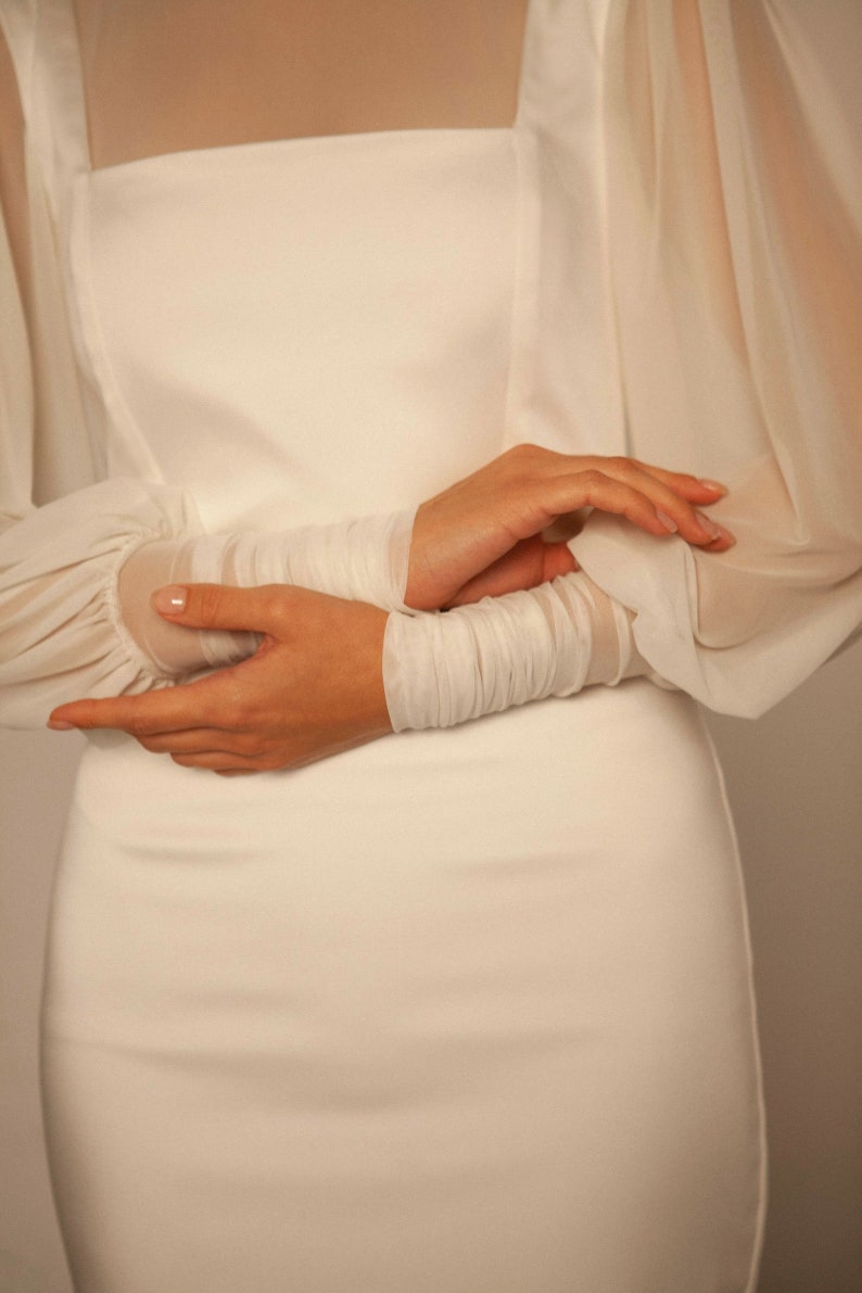 May include: A white dress with long sleeves and a fitted bodice. The sleeves are made of sheer fabric and have a gathered detail at the wrist.