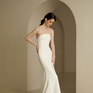 Puede incluir: Un sencillo vestido de novia blanco sin tirantes, con un corpi&ntilde;o ajustado y una falda fluida.