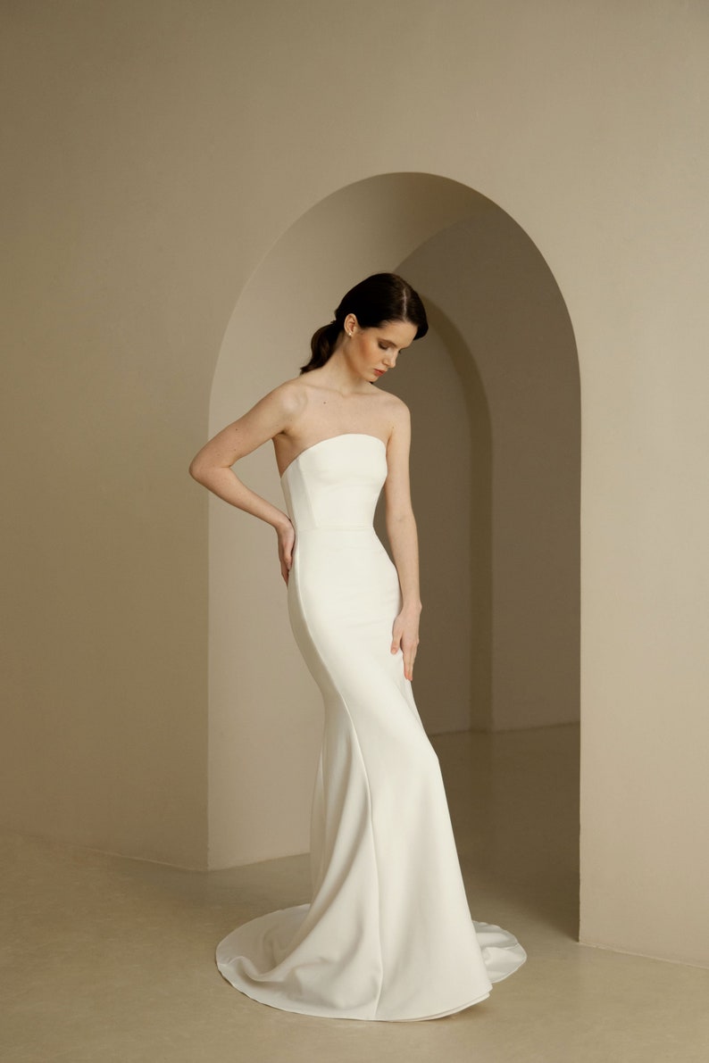 Puede incluir: Un sencillo vestido de novia blanco sin tirantes, con un corpi&ntilde;o ajustado y una falda fluida.