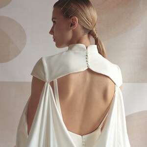 Puede incluir: Un vestido blanco de sat&eacute;n con la espalda descubierta y un cuello alto. El vestido tiene un dise&ntilde;o de capa en la espalda y se abrocha con botones.