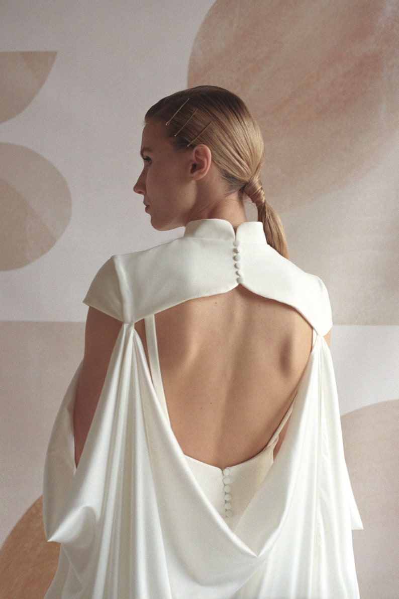 Puede incluir: Un vestido blanco de sat&eacute;n con la espalda descubierta y un cuello alto. El vestido tiene un dise&ntilde;o de capa en la espalda y se abrocha con botones.