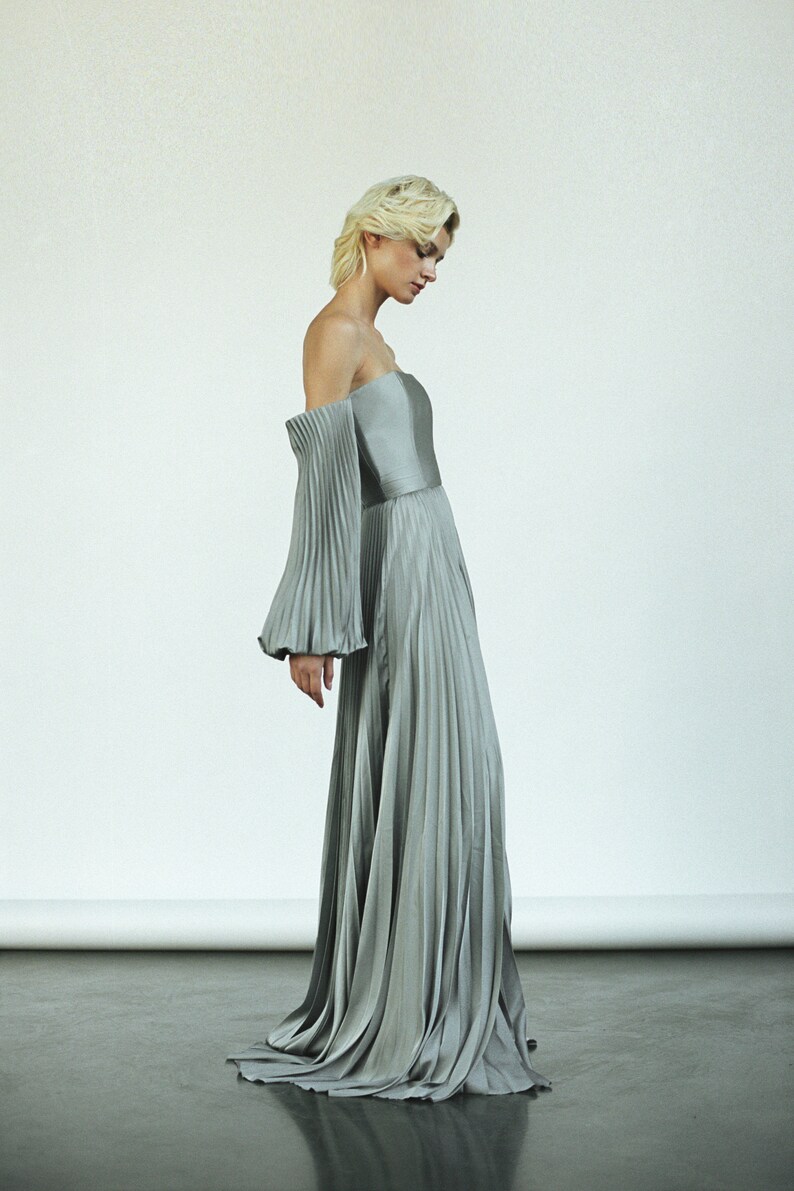May include: A silver off-the-shoulder gown with pleated fabric and long sleeves.