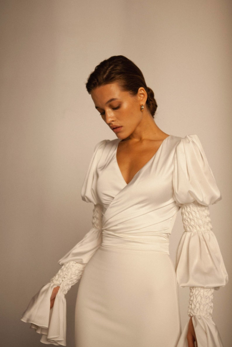 May include: A white satin wrap dress with long sleeves and a V-neckline. The sleeves have large, gathered cuffs.
