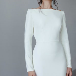 May include: A white long-sleeved dress with a bateau neckline and a fitted silhouette. The dress is made of a smooth, matte fabric.