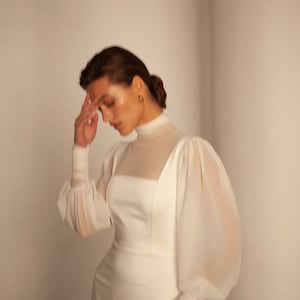 May include: A white dress with a high neck and long sleeves. The sleeves are puffed and made of a sheer fabric. The dress is fitted and has a simple design.
