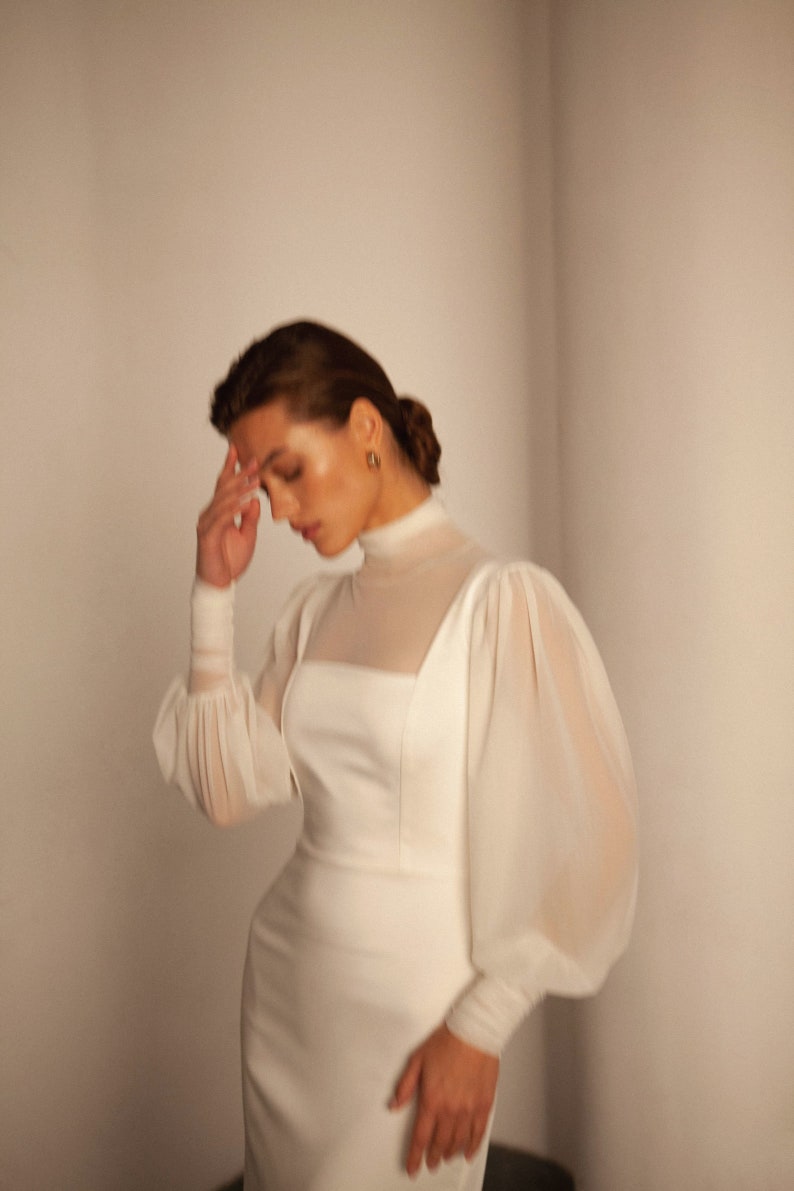 May include: A white dress with a high neck and long sleeves. The sleeves are puffed and made of a sheer fabric. The dress is fitted and has a simple design.