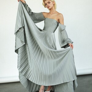 May include: A woman wearing a light gray pleated dress with off-the-shoulder sleeves and white heels.