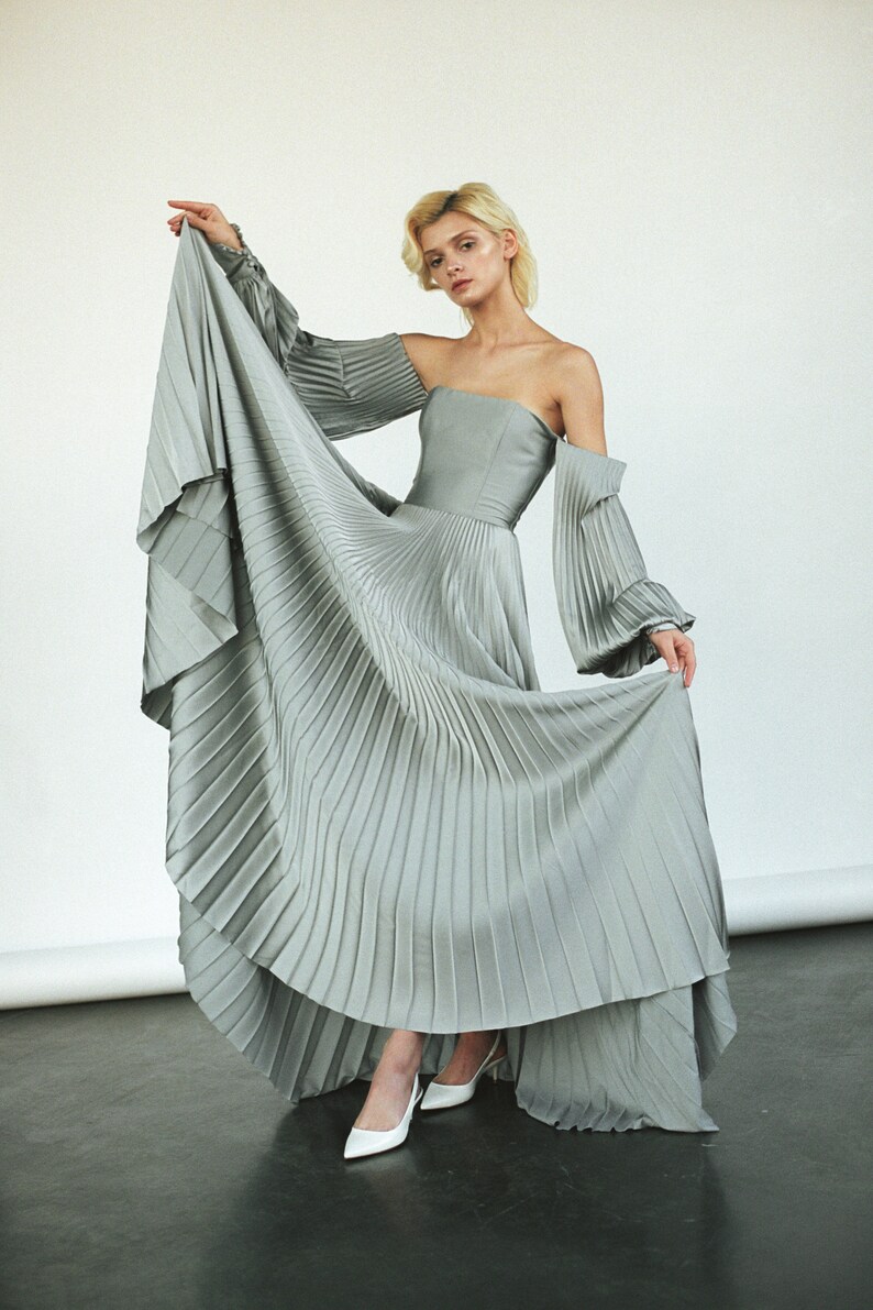 May include: A woman wearing a light gray pleated dress with off-the-shoulder sleeves and white heels.