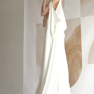 Puede incluir: Un vestido blanco sin mangas con cuello alto y falda fluida. El vestido est&aacute; drapeado y tiene un dise&ntilde;o &uacute;nico. La modelo est&aacute; de pie frente a una pared con un patr&oacute;n geom&eacute;trico.
