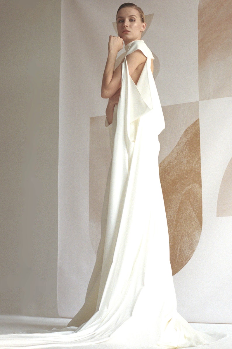 Puede incluir: Un vestido blanco sin mangas con cuello alto y falda fluida. El vestido est&aacute; drapeado y tiene un dise&ntilde;o &uacute;nico. La modelo est&aacute; de pie frente a una pared con un patr&oacute;n geom&eacute;trico.