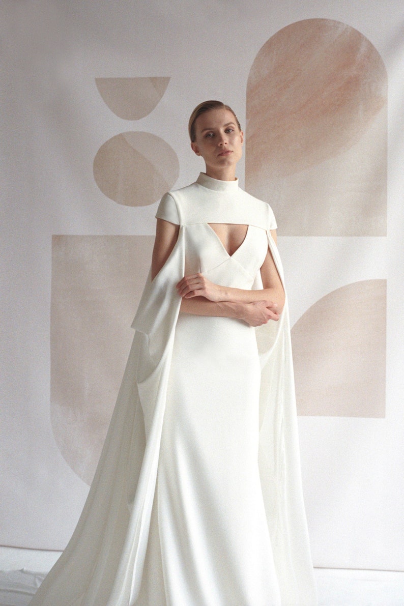 Puede incluir: Un vestido de novia blanco con cuello alto y una larga cola. El vestido tiene un corpi&ntilde;o ajustado y una falda fluida. La modelo est&aacute; de pie frente a un fondo blanco con formas abstractas.