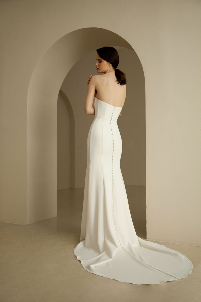Puede incluir: Un vestido de novia blanco sin tirantes con un corpi&ntilde;o ajustado y una falda fluida. El vestido tiene una cola larga y una fila de botones en la espalda.