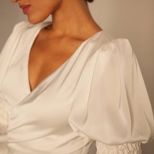 May include: A white satin dress with a v-neckline and long sleeves. The sleeves are puffed at the shoulders and have a gathered detail at the cuffs.