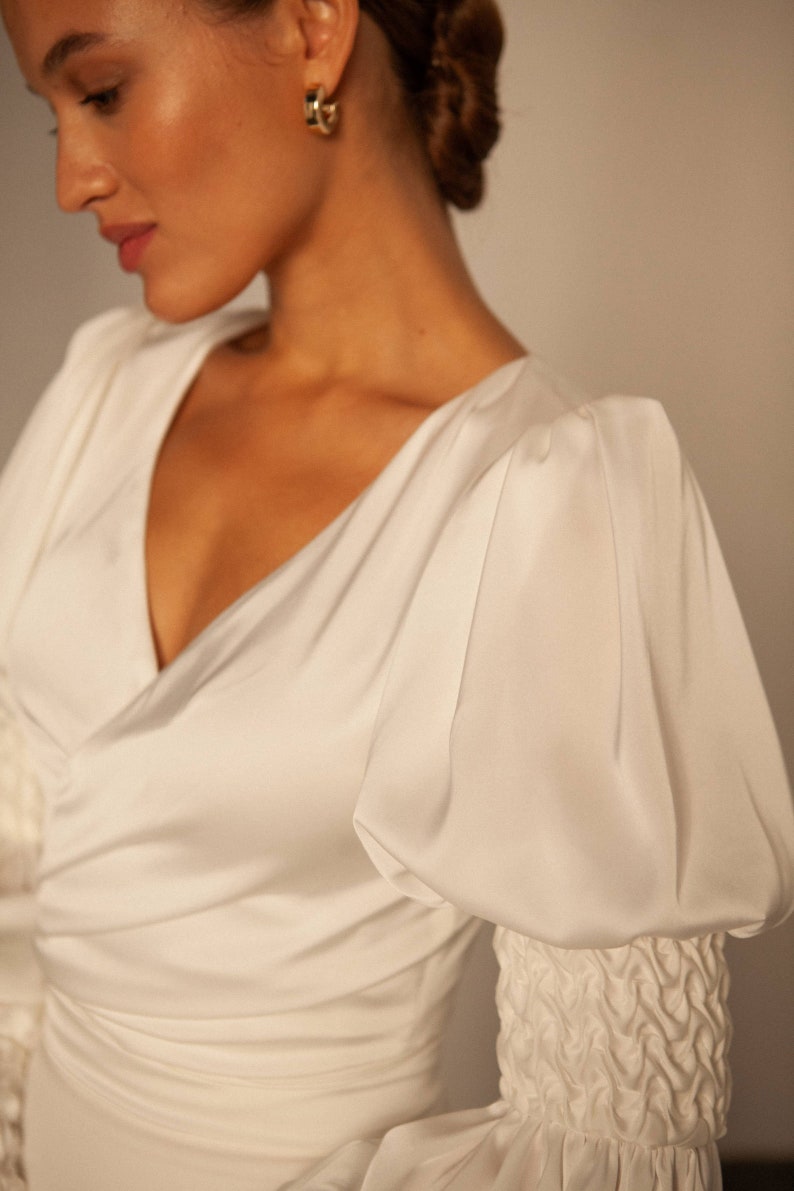 May include: A white satin dress with a v-neckline and long sleeves. The sleeves are puffed at the shoulders and have a gathered detail at the cuffs.