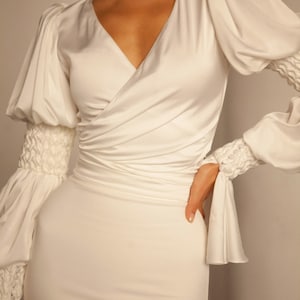 May include: A white satin dress with a V-neckline and long sleeves. The sleeves have a gathered detail at the cuffs. The dress is fitted and has a wrap-style bodice.
